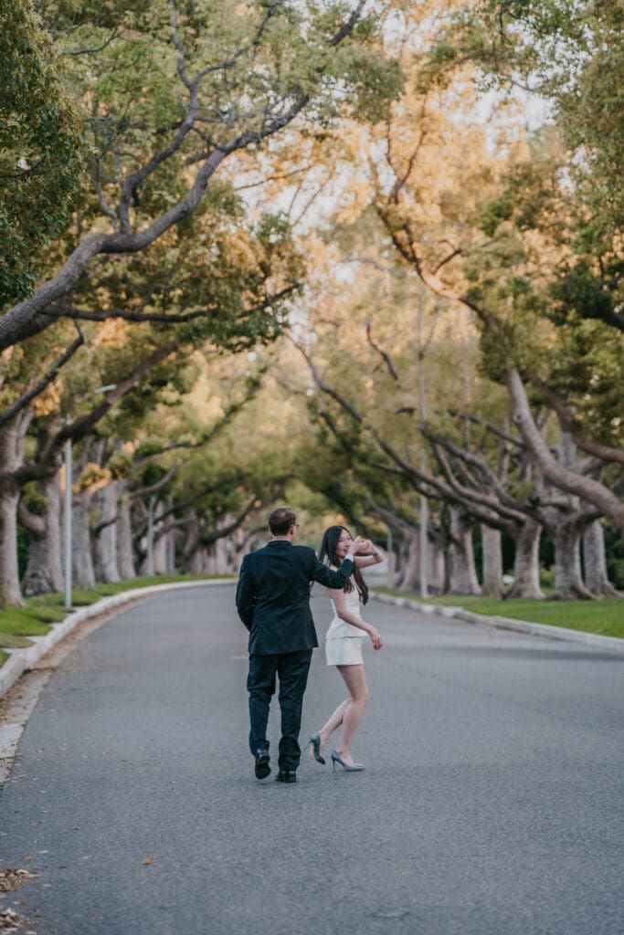 Couple walking hand in hand beneath mature trees on a Beverly Hills street during engagement photos