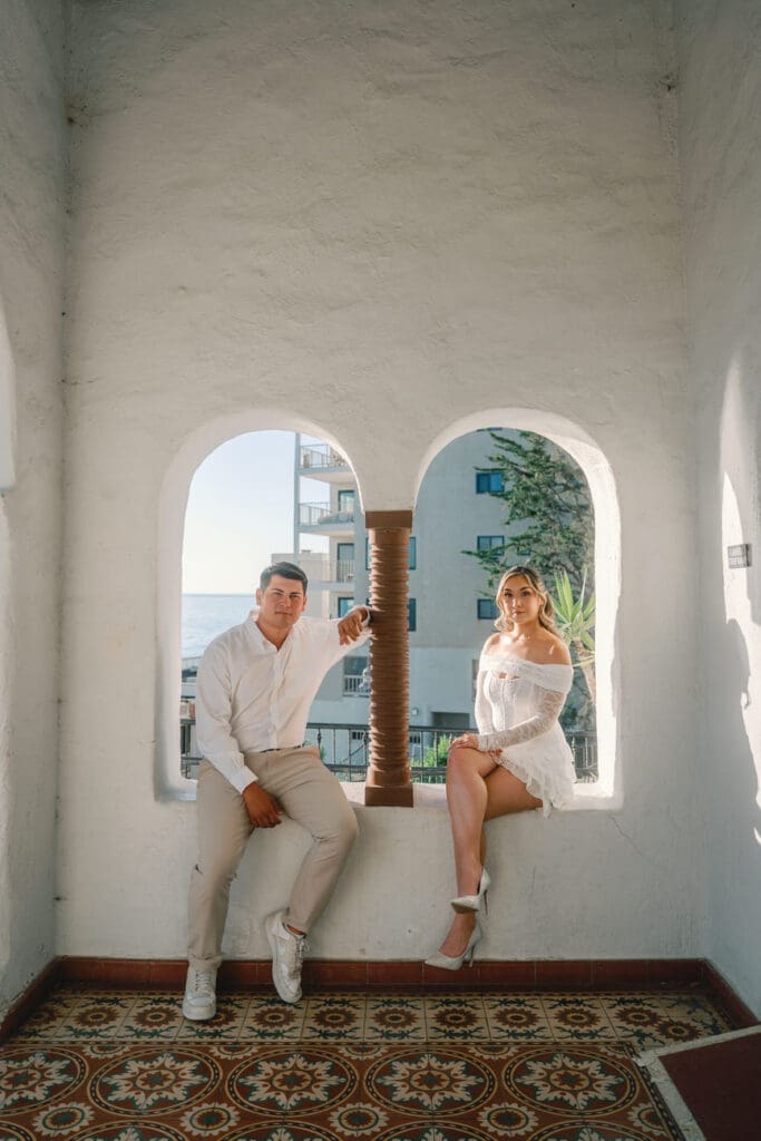 Couple sitting together near the arched windows at Casa Romantica during their San Clemente engagement photos.