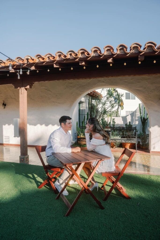 Couple seated together at a small courtyard table during their Casa Romantica engagement session in San Clemente.