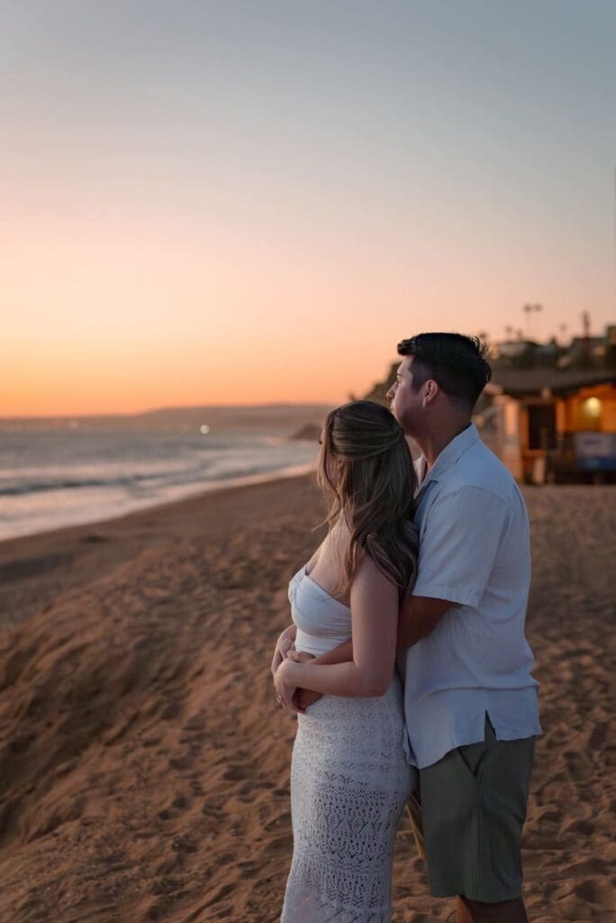 Couple standing near San Clemente Beach at sunset with the pier in the background after their Casa Romantica engagement photos.
