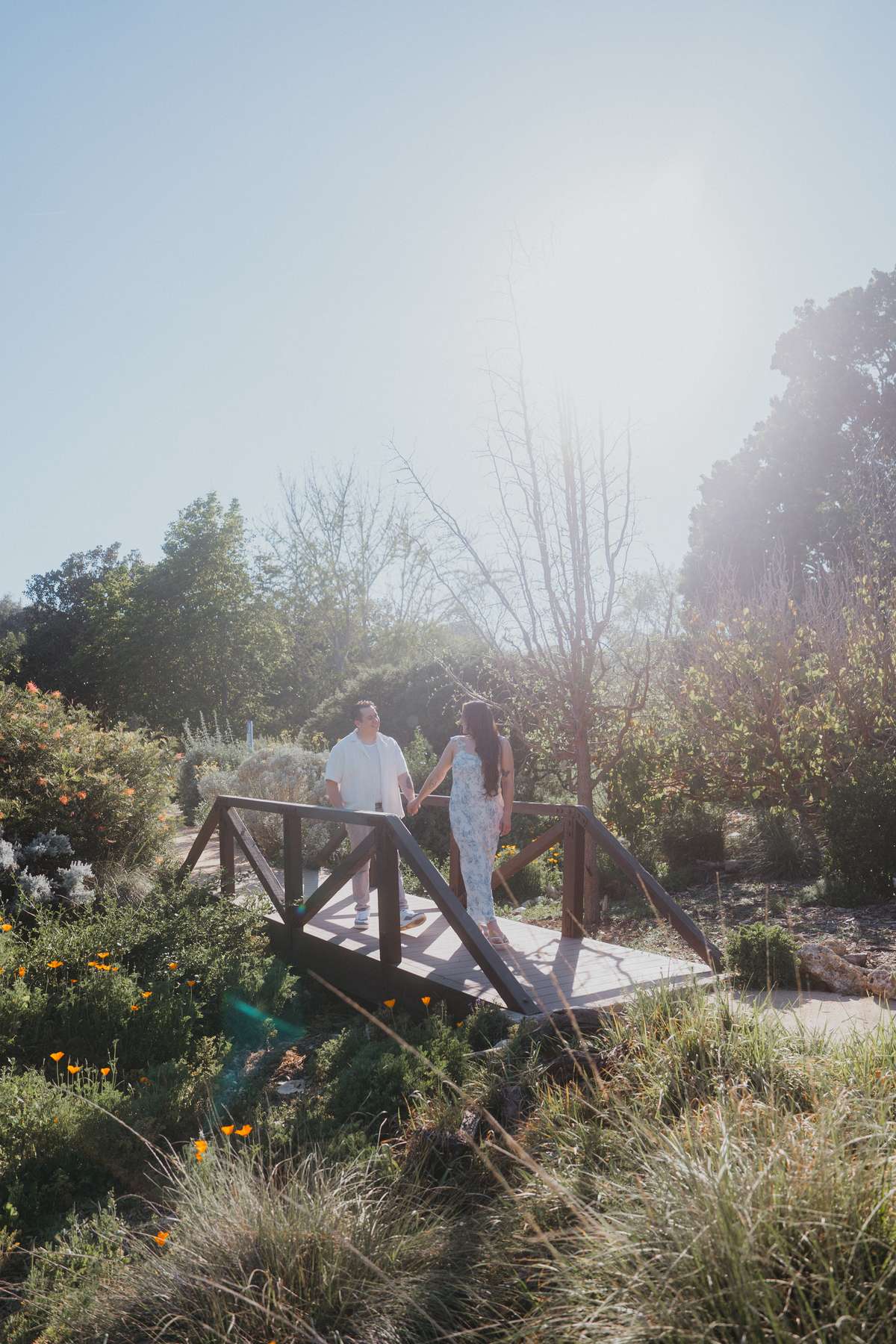 Couple walking together across a small garden footbridge in bright afternoon light.