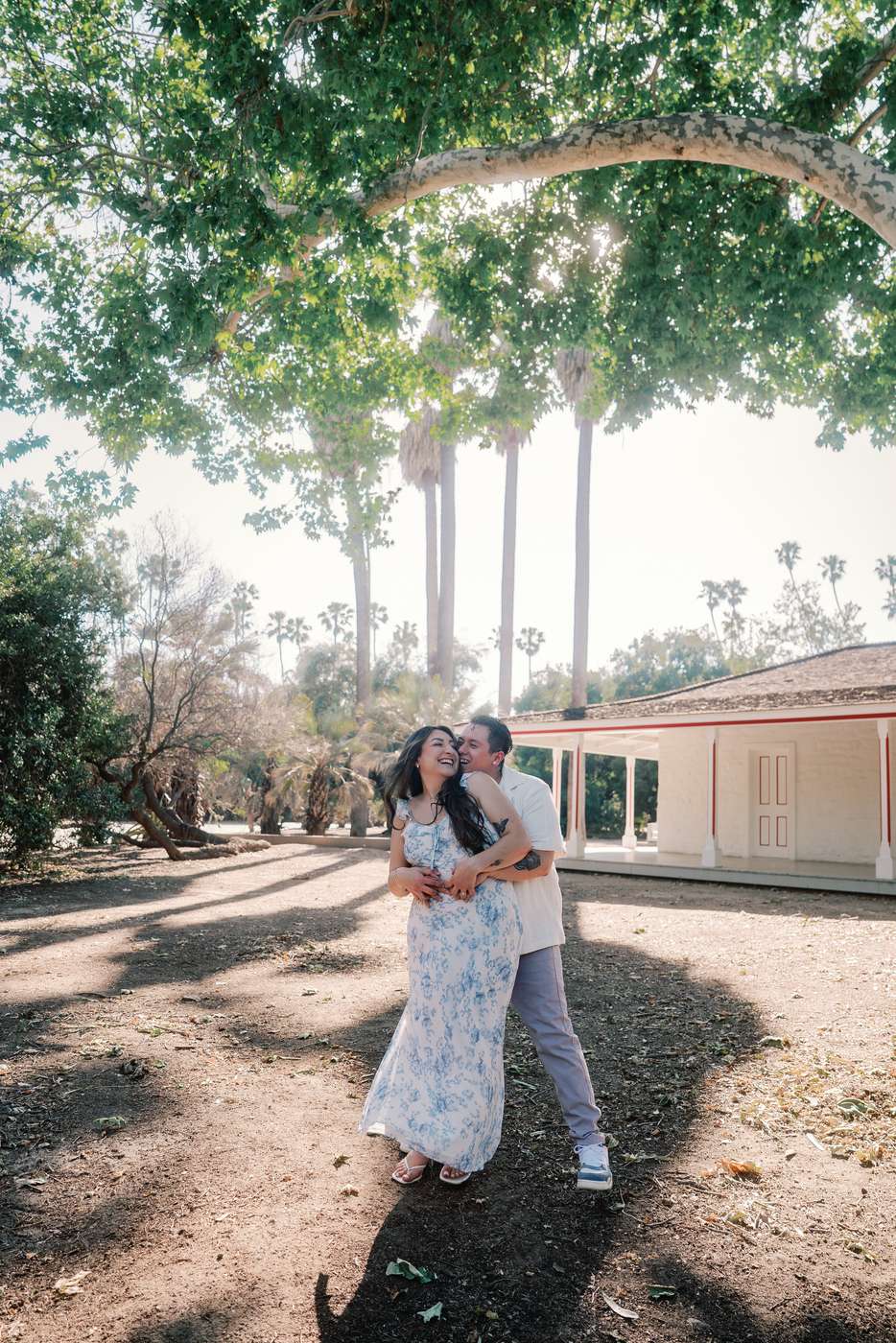 Couple standing beneath a broad tree canopy beside a historic red-trimmed pavilion.