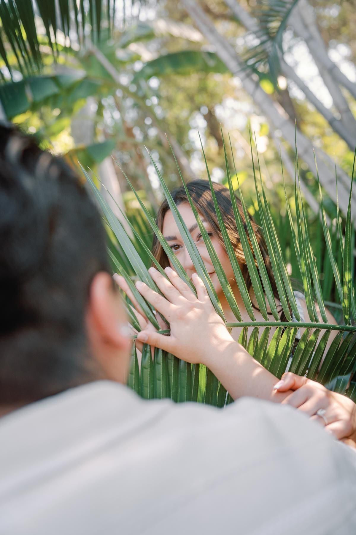 Woman smiling through palm leaves during a playful engagement portrait.