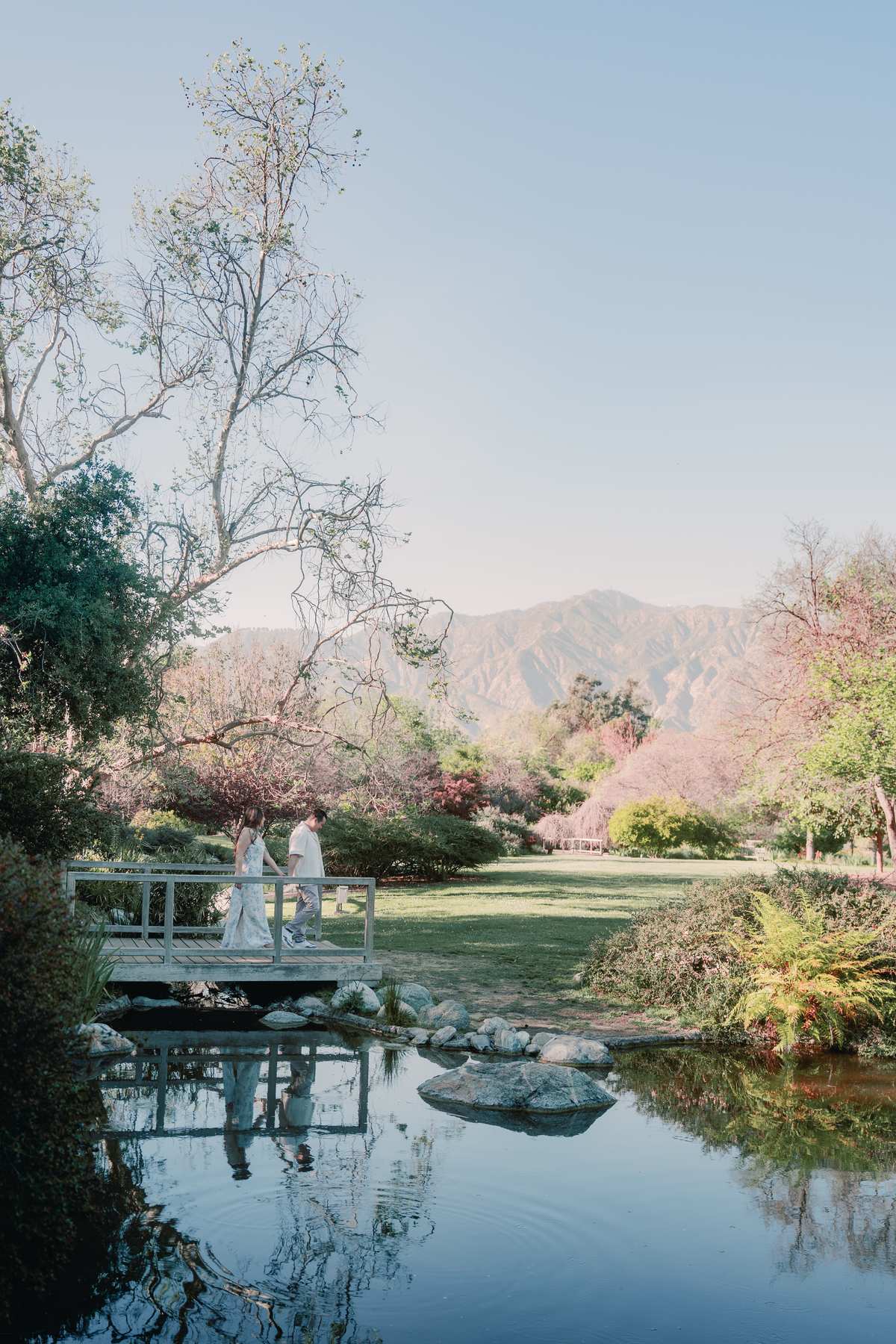 Couple crossing a white footbridge reflected in a pond with mountain views beyond.