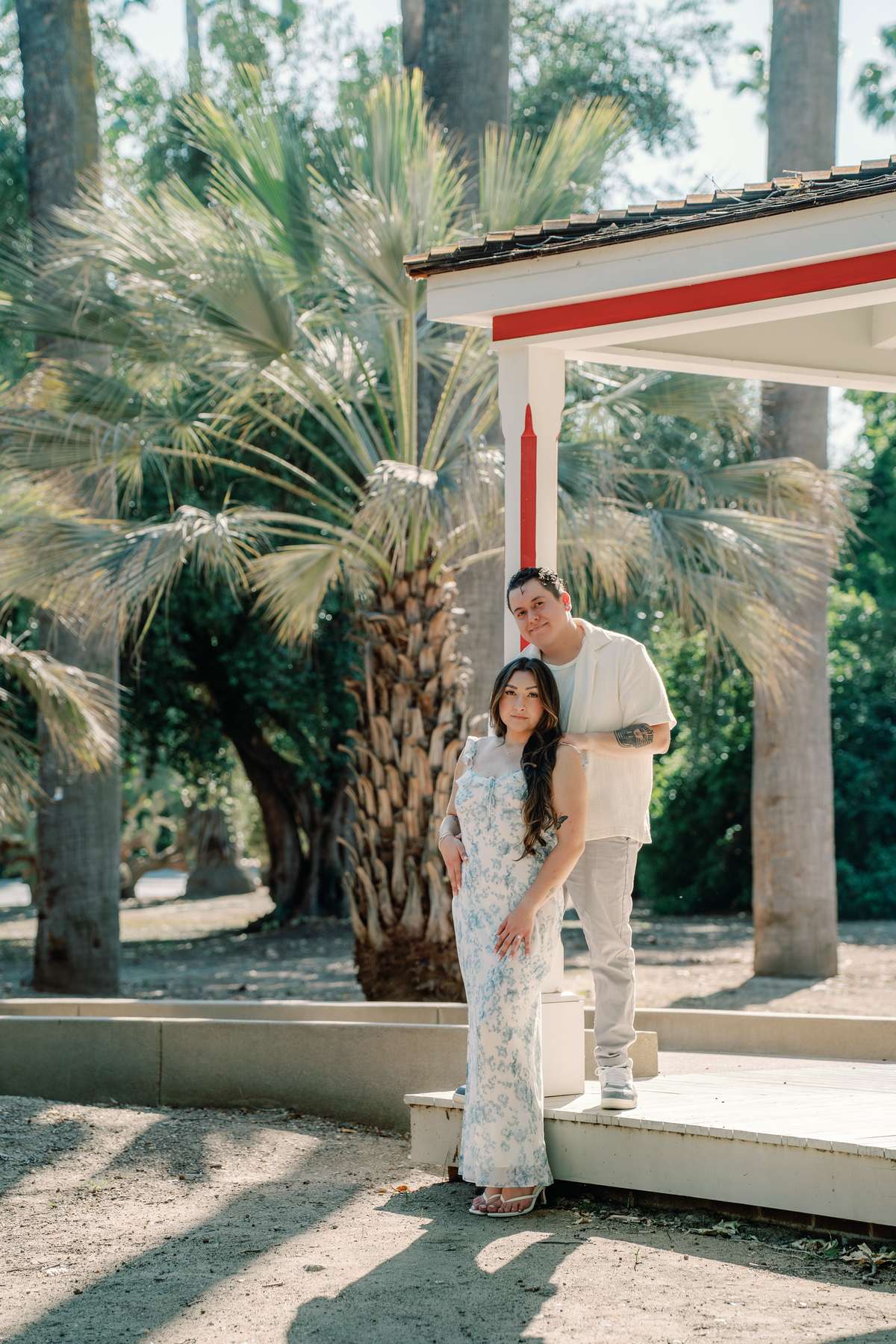 Couple posing beside a red-trimmed porch and palms at the LA Arboretum.