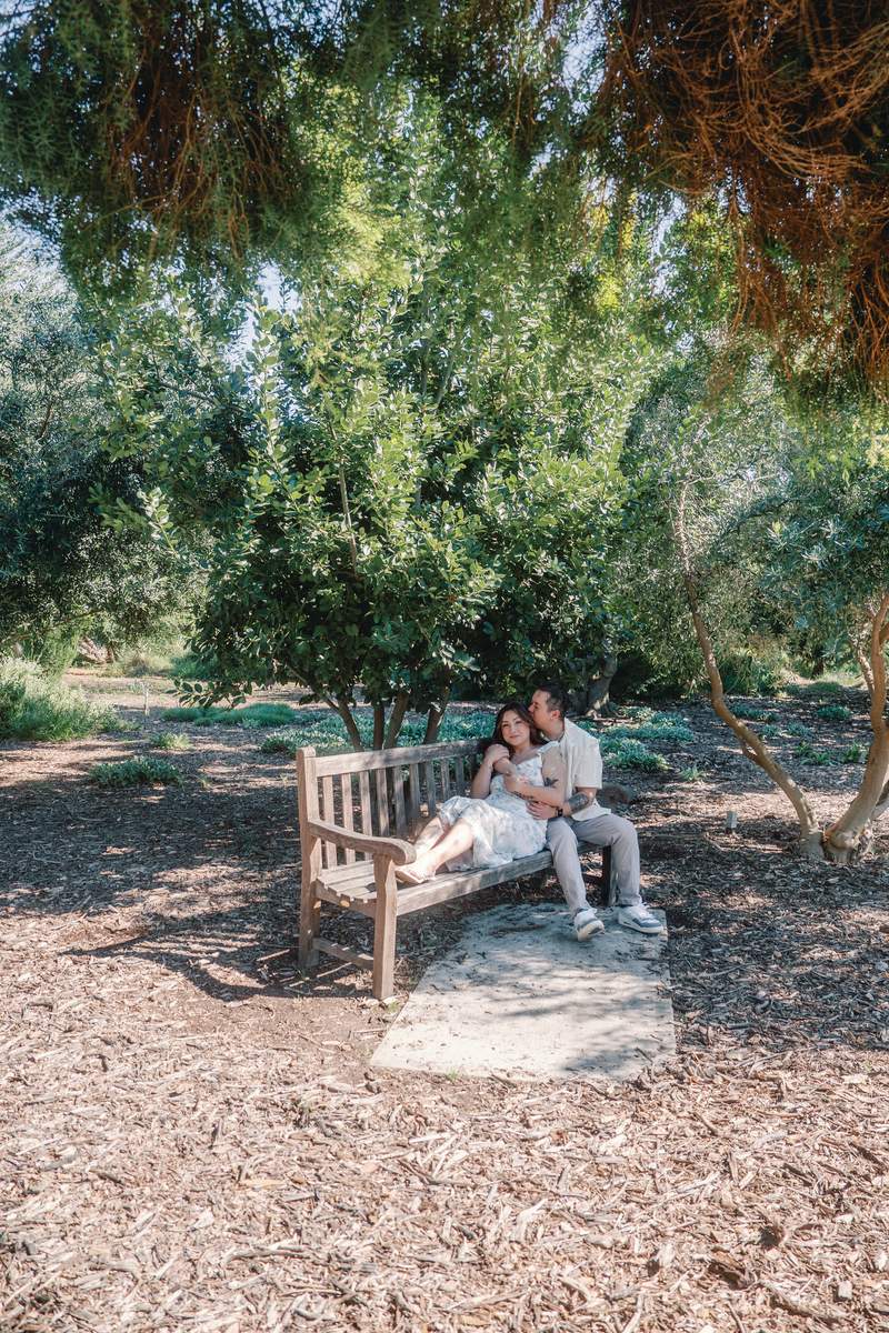 Couple sitting close together on a shaded wooden bench beneath dense trees.