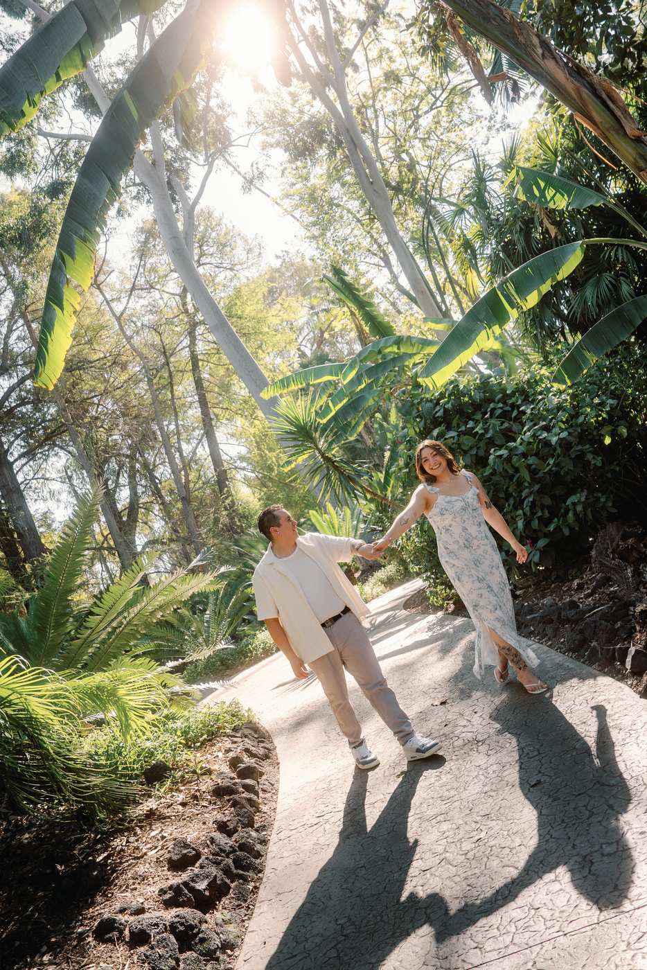 Couple holding hands while walking along a sunlit tropical garden path.