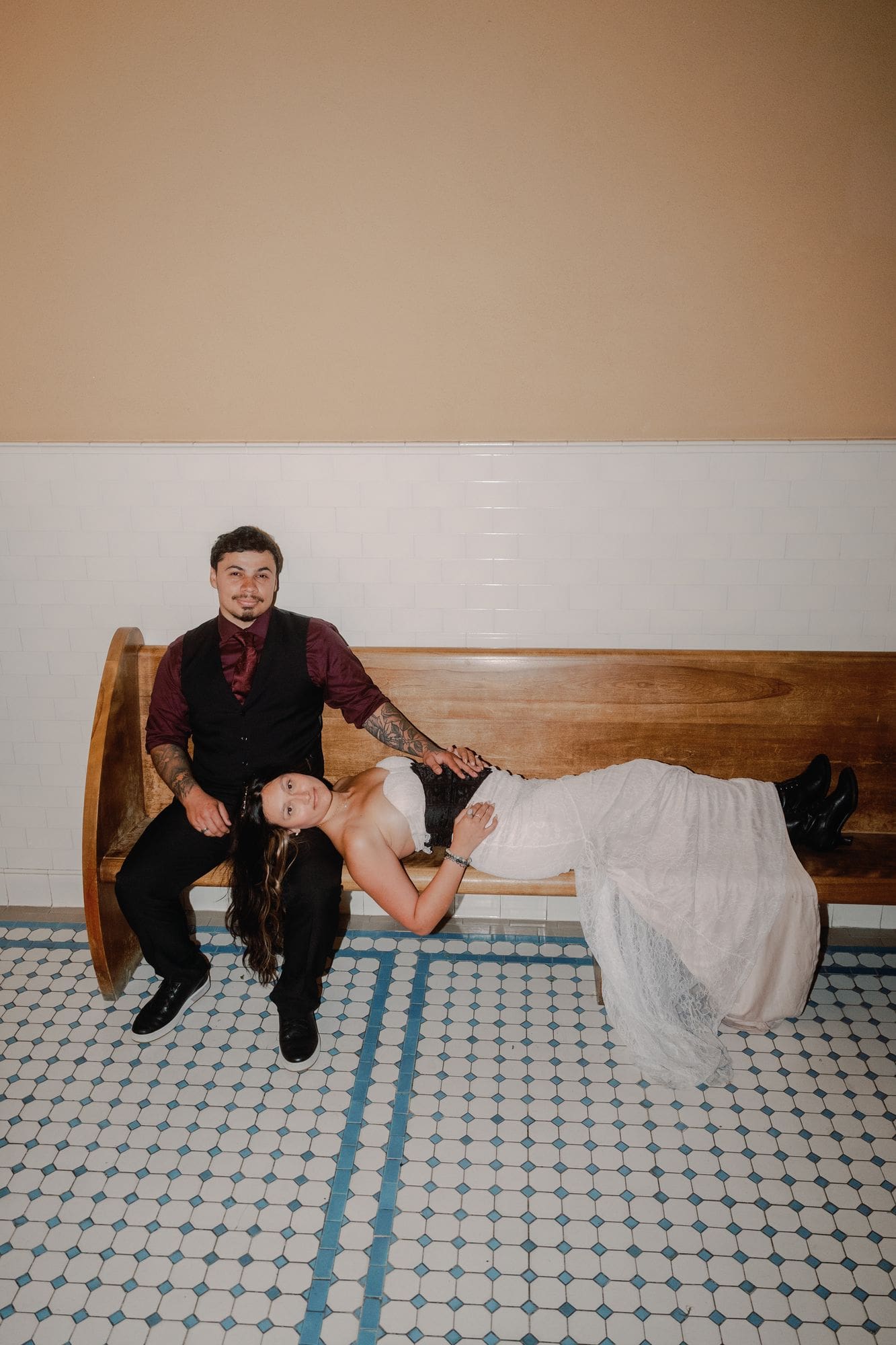 The bride reclines across her partner’s lap on a wooden bench.