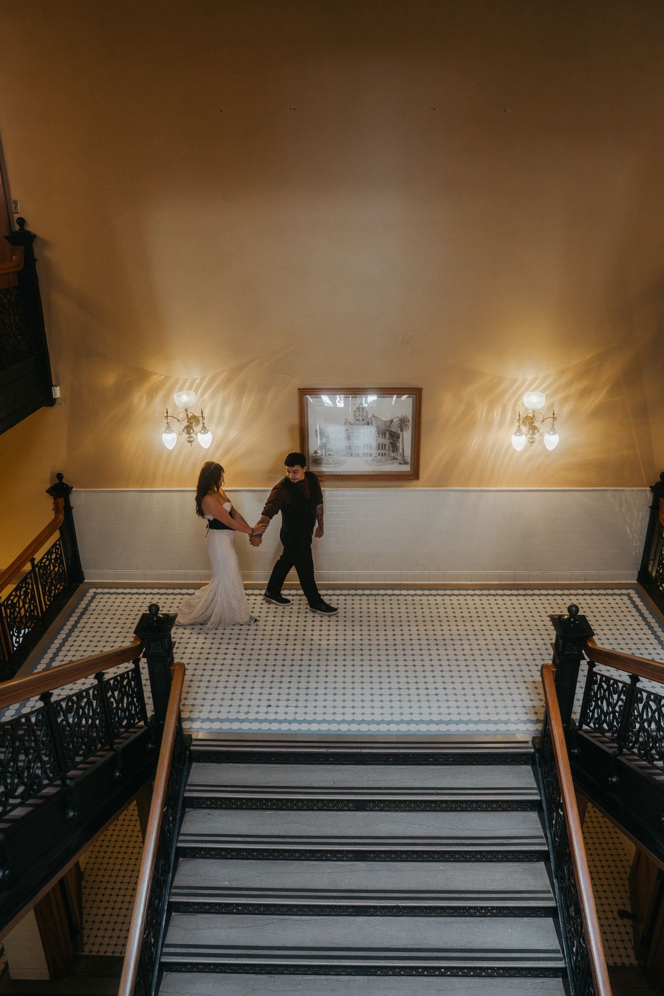 The couple holds hands on a tiled landing beneath the staircase.