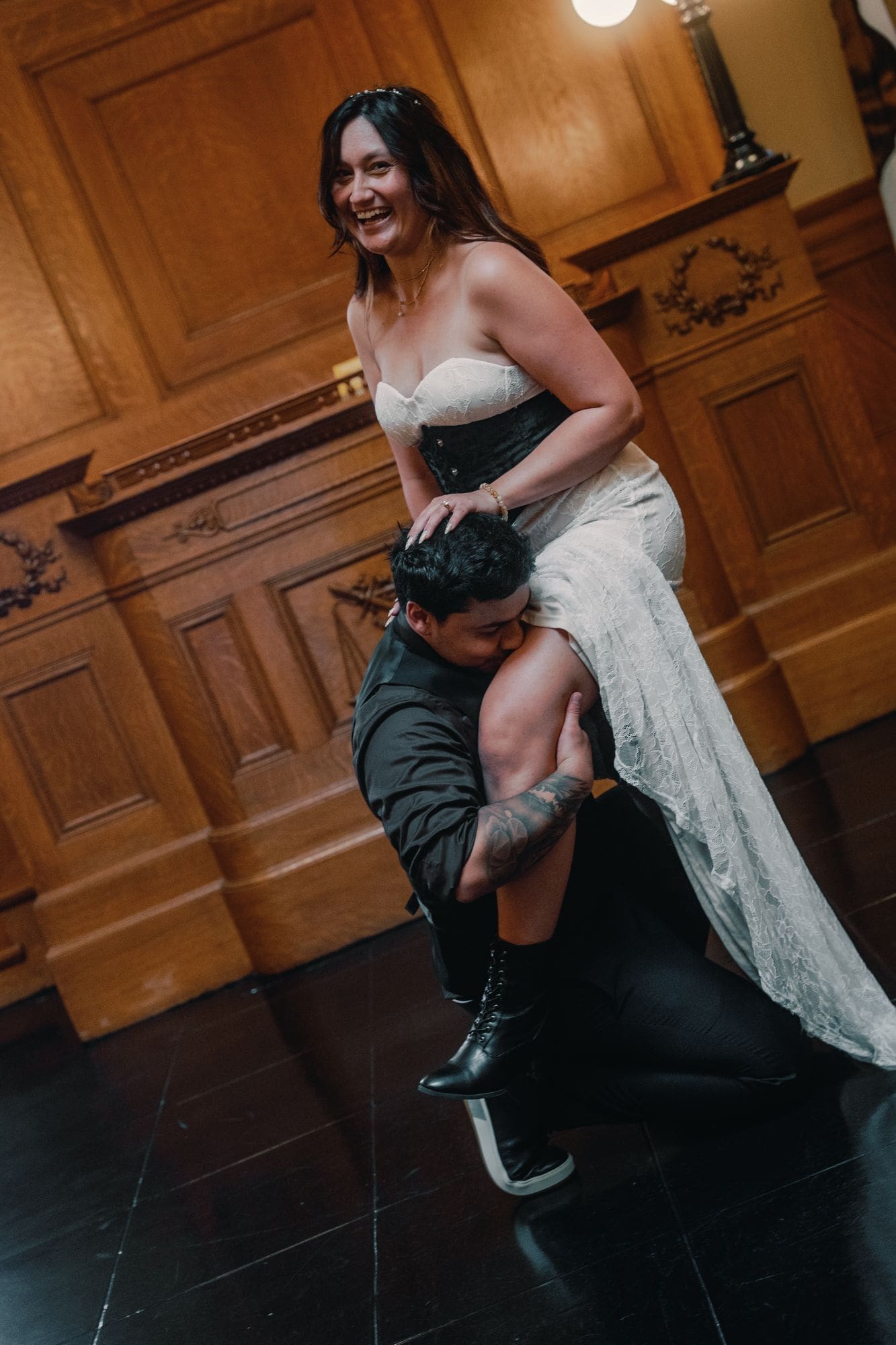 The bride laughs while lifted over her partner’s shoulder in a wood-paneled room.