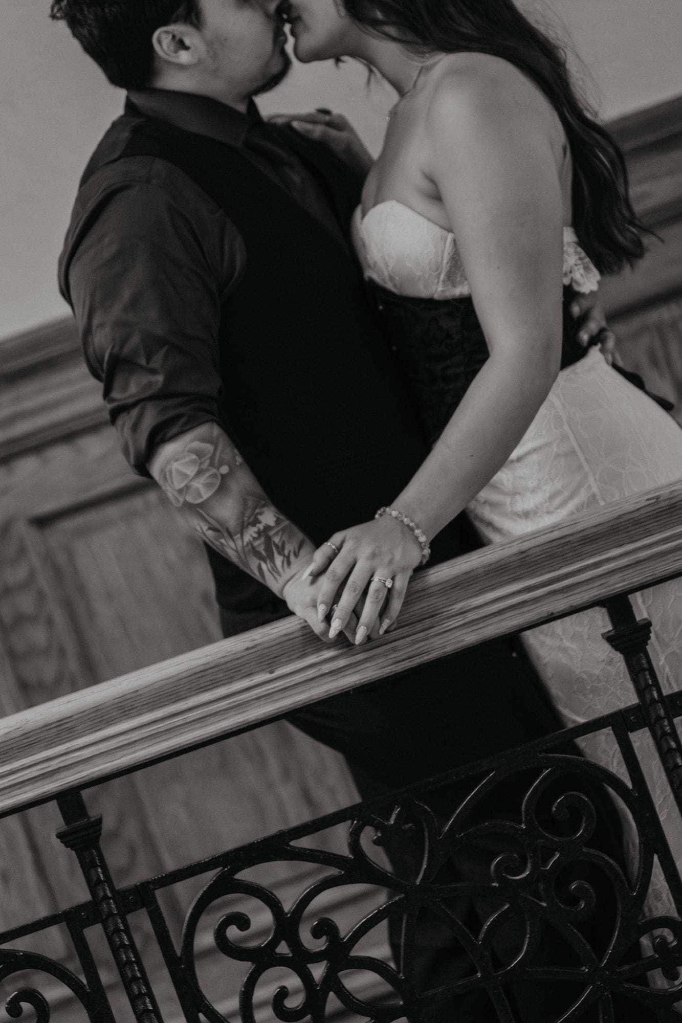 A black-and-white portrait of the couple kissing beside an ornate railing.