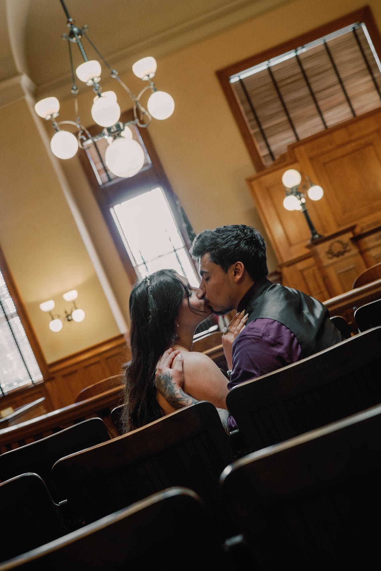 The couple kisses while seated among dark wooden chairs.