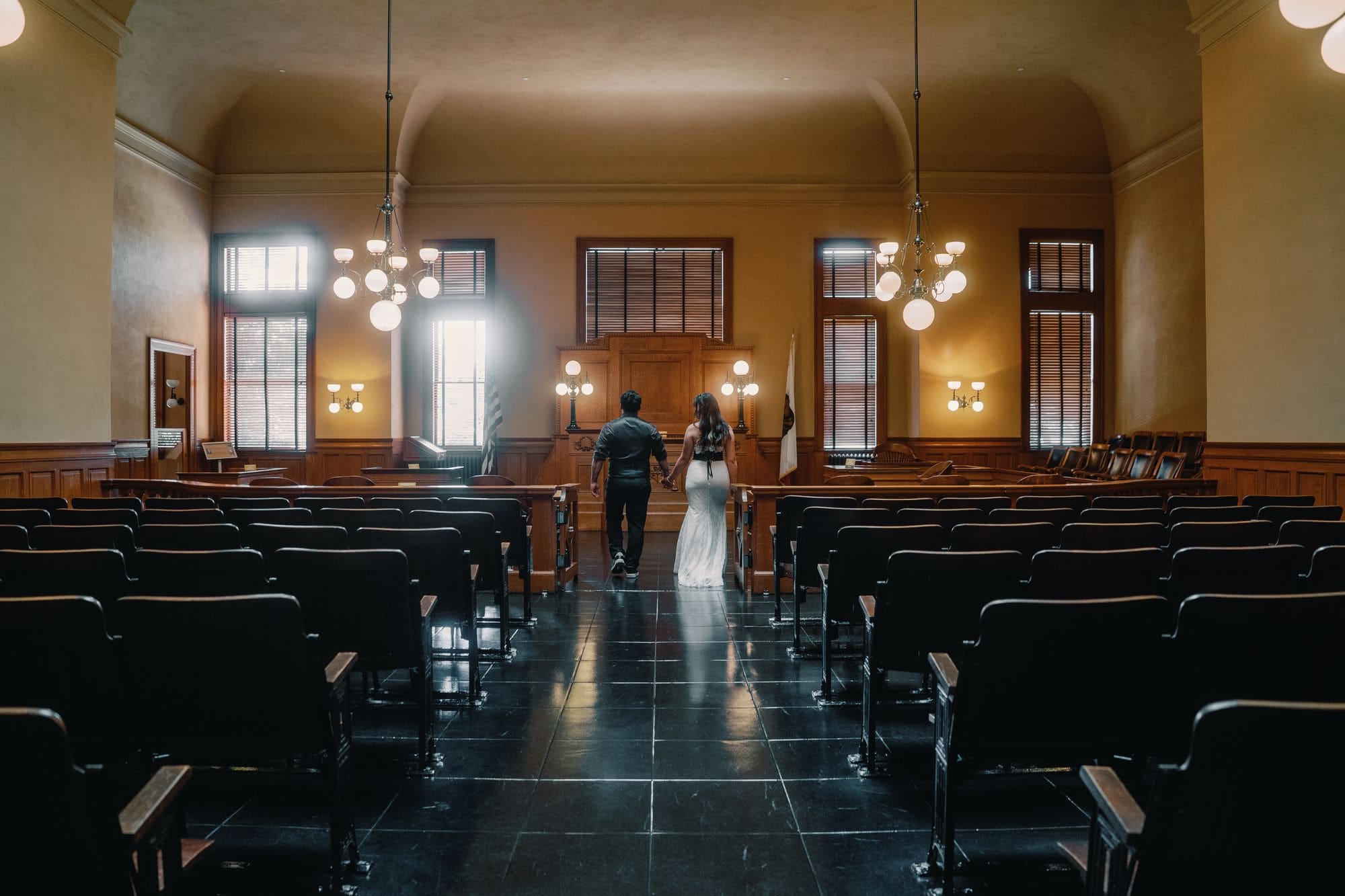 The couple walks between rows of chairs in an empty wood-paneled room during their Santa Ana courthouse wedding portraits.