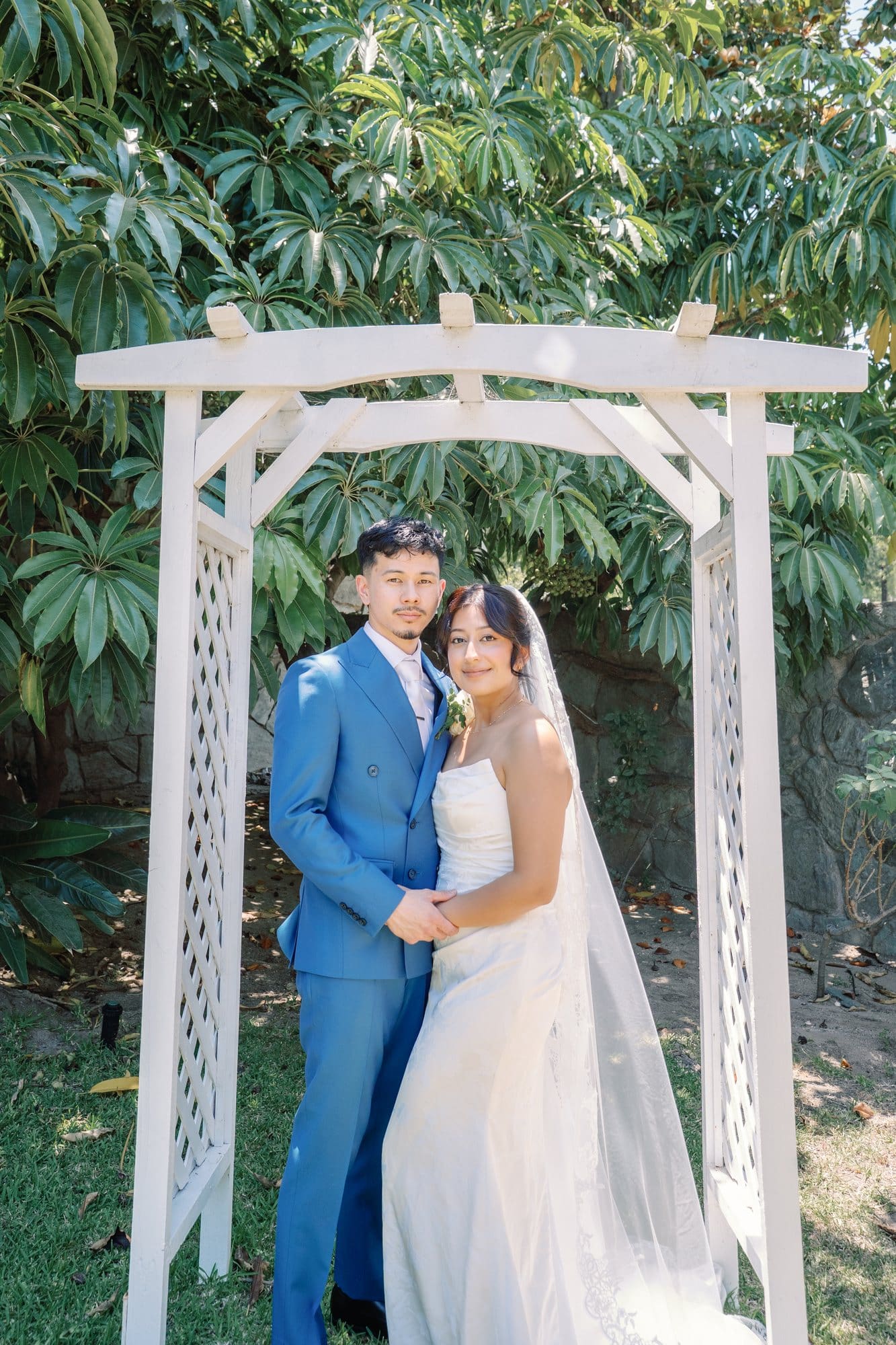 The couple stands together under a white garden arbor at The Santa Anita Church in Arcadia, California.