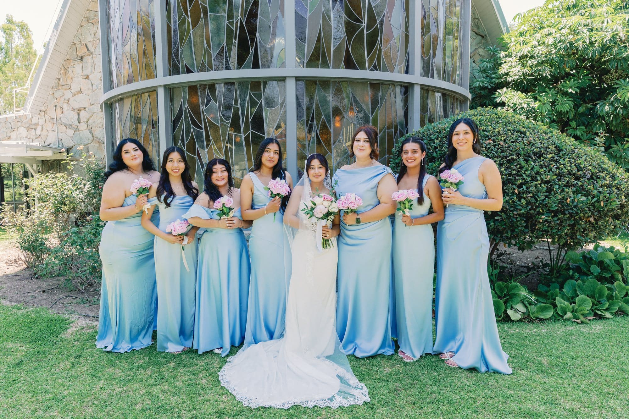 The bride stands with bridesmaids in pale blue dresses beside the tower at The Santa Anita Church in Arcadia, California.