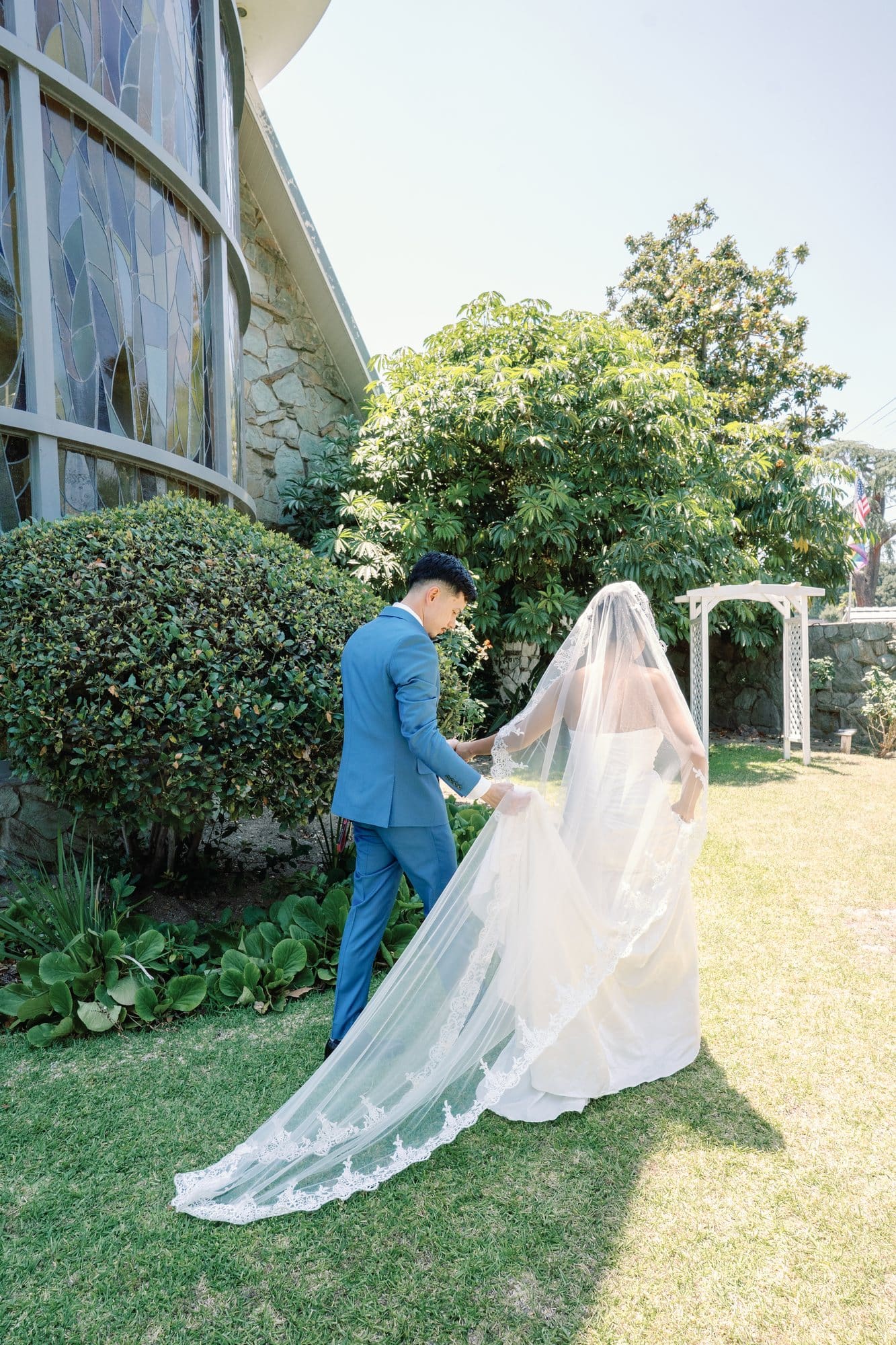 The couple walks hand in hand through the garden with the bride’s veil trailing behind at The Santa Anita Church in Arcadia, California.
