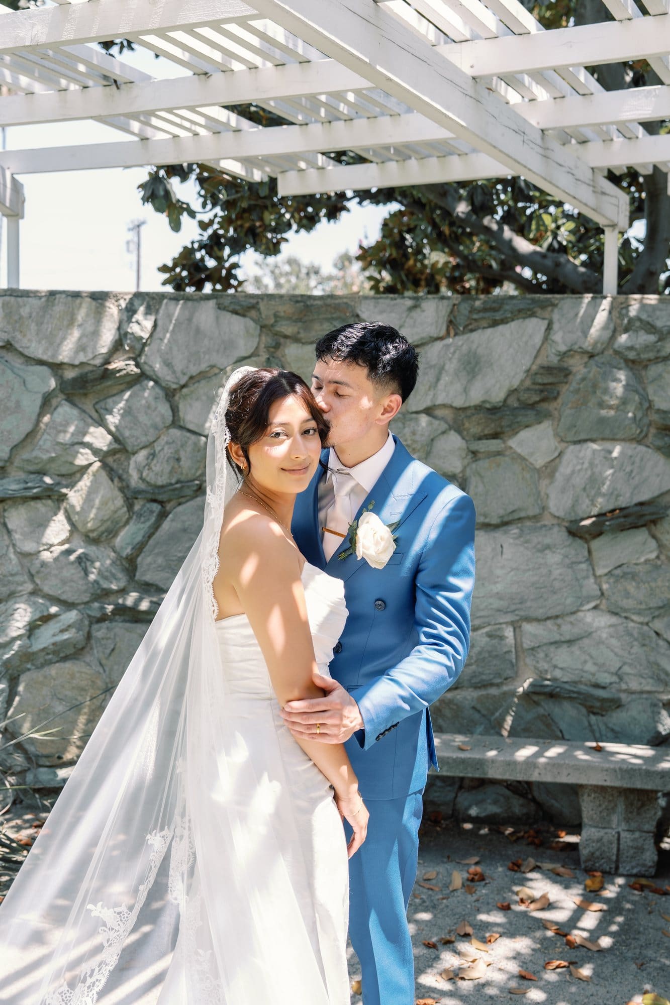 The couple embraces beneath a white pergola beside a stone wall at The Santa Anita Church in Arcadia, California.